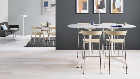 Cuatro Taburetes Leeway con bases metálicas y asientos y respaldos de madera clara, junto a una mesa alta cerca de una tumbona con un sofá negro y sillas alrededor de una mesa.