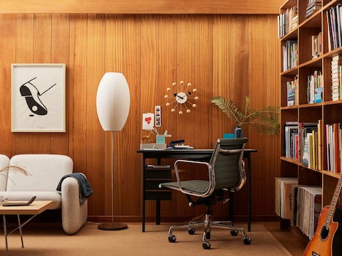 Escritorio Mode en negro con superficie en sandstone en una sala de biblioteca con paneles de madera, lámpara de cigarro y sofá Wilkes en crema.