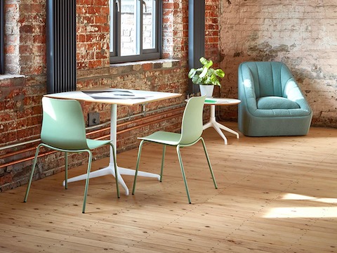An Ali Café Table with a white 4-star base and square top, with two Polly Side Chairs around it. A blue Fiji Chair is in the background of the setting.