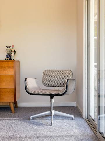 Una sola silla tapizada en blanco y negro con patas de metal se encuentra en un rincón de una habitación, junto a puertas corredizas y una pequeña cómoda de madera.
